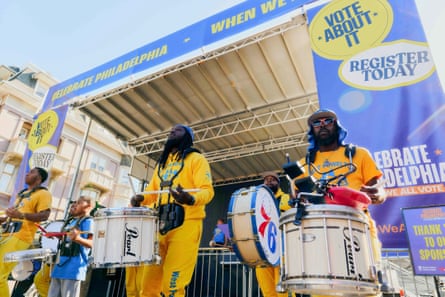people in yellow and blue outfits stand outside playing drums next to banner about voting