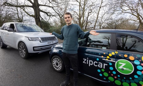 Jasper Jolly stands in the street in front of a black Zipcar-branded Fiat 500 with his hands outstretched; a silver Range Rover is parked nose-on to the Fiat.