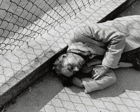 A person lies on pavement looking up, with shadow patterns cast across their body and the ground from a chain link fence