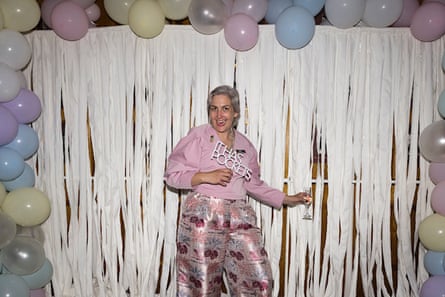 A woman in a pink blouse with pleated sleeves and metallic jacquard pants, standing against a white fringe curtain framed by pastel-coloured balloons.