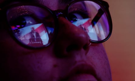 A tight closeup of a young woman’s face, the lights of the stage reflected in her round glasses.