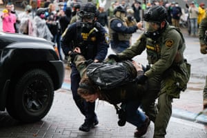 Woman with arms twisted back being forced forward by two officers wearing gas masks