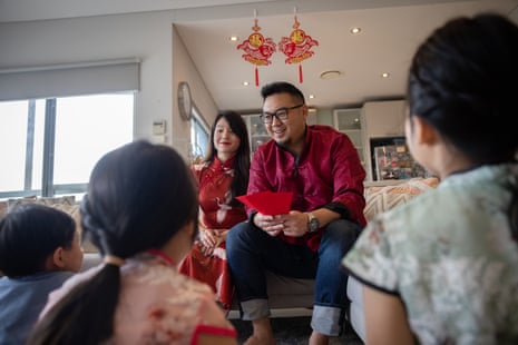 A mother and father sitting on a couch holding red packets of money for lunar new year; their three children sit before them on the floor