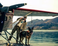 Brad Pitt stands on a floatplane's pontoon beside a German shepherd, lake and mountains behind