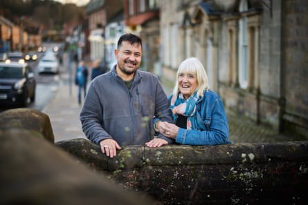 Samir and Sue stand behind a stone wall in a street of stone buildings, both smiling; he appears to be in his late 30s, has short dark hair and wears a grey fleece jumper, and she is older, with long blond hair cut in a fringe, and wears a denim jacket and patterned scarf
