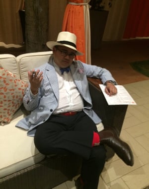 A man in a hat, bow tie, striped jacket and red socks, sits cross legged on a sofa, holding a cigar
