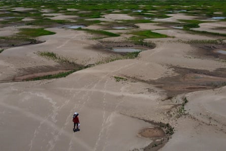 A person walks over mud flats with a bag of provisions on their head.