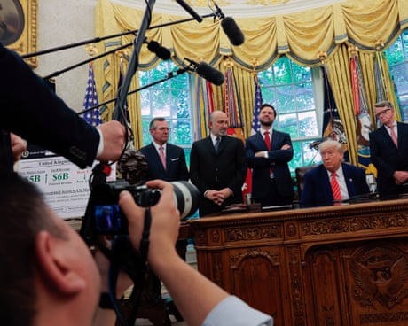a man seated a desk surrounded by people talks to a group holding cameras and microphones
