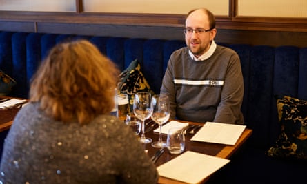 A young, balding man with glasses, wearing a grey top over a white shirt, and an older woman with curly red hair and glasses, wearing a grey sparkly top, sitting at a wooden restaurant table talking