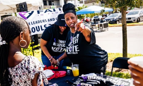 Three Black women in black T-shirts stand and gesture near a foldout table outside.