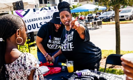 On a stretch of grassy lawn beside a small parking lot, two Black women stand behind a table with stacks of folded T-shirts - which they also wear - that say 'Power, Respect, Let's Vote' in black, purple and white.