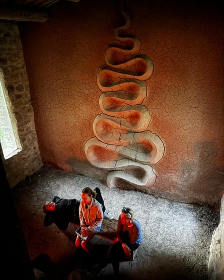 Three women sit on a bench in a simple stone building. Behind them a serpentine shape made of dried mud snakes up the wall.