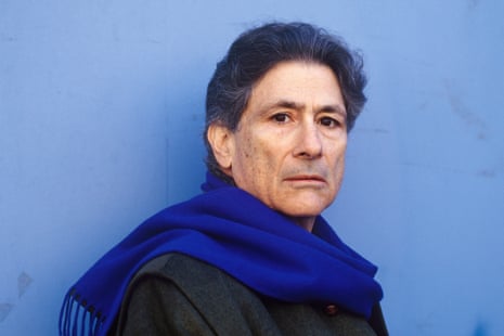 A man in a blue scarf against a blue background looks  into the camera.