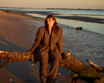 The musician Wendy Eisenberg wearing a suit and sitting on a tree trunk on a beach