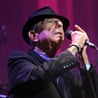 Portrait of Leonard Cohen in a suit and fedora, singing into the mic, looking upwards