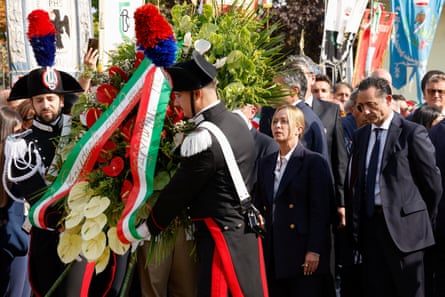 Giorgi Meloni stands at the front of a group of people as a soldier in ceremonial uniform with tall feathered hat lays a large wreath of flowers bedecked with red, white and green ribbon. She has her long blond hair tied back and stands with her head held high, surrounded by men who are much taller than her.