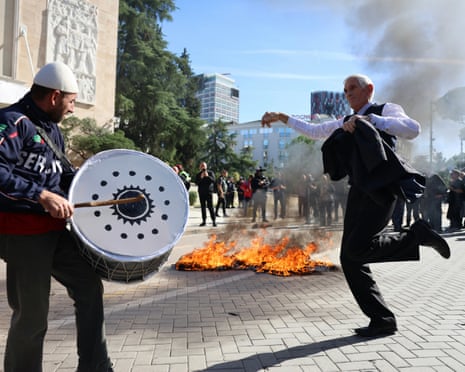 A man dancing while another man plays a drum, with fire burning in the street behind them