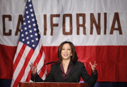 woman who is Kamala Harris wears black suit and gestures behind podium and microphone with US flag behind her
