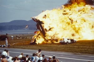 A crashed jet in flames by a runway as people look on.