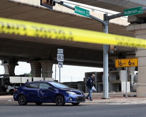 dark blue toyota sedan dented with apparent bullet holes as a police officer stands on sidewalk nearby