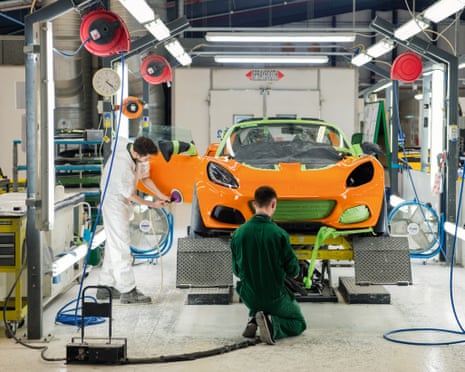 Lotus cars assembly line: two men in overalls are working on a bright orange sports car on a ramp inside a manufacturing facility.