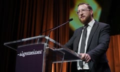 Dovid Efune speaking at a podium table branded with the name of The Algemeiner