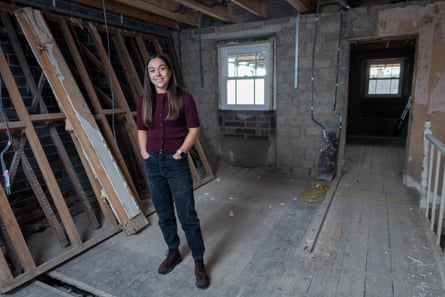 Alice Helps stands in a very unfinished first-floor room with bare brick and no ceilings