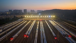 An aerial shot of a train station with a row of trains, floodlights and a cityscape in the background