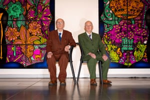 The artists Gilbert and George sitting in front of some of their brightly coloured work