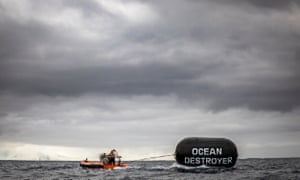 A stormy sky and a rubber dingy at sea (with a few people on it) towing a large ship's fender with the words ‘Ocean Destroyer’ on it