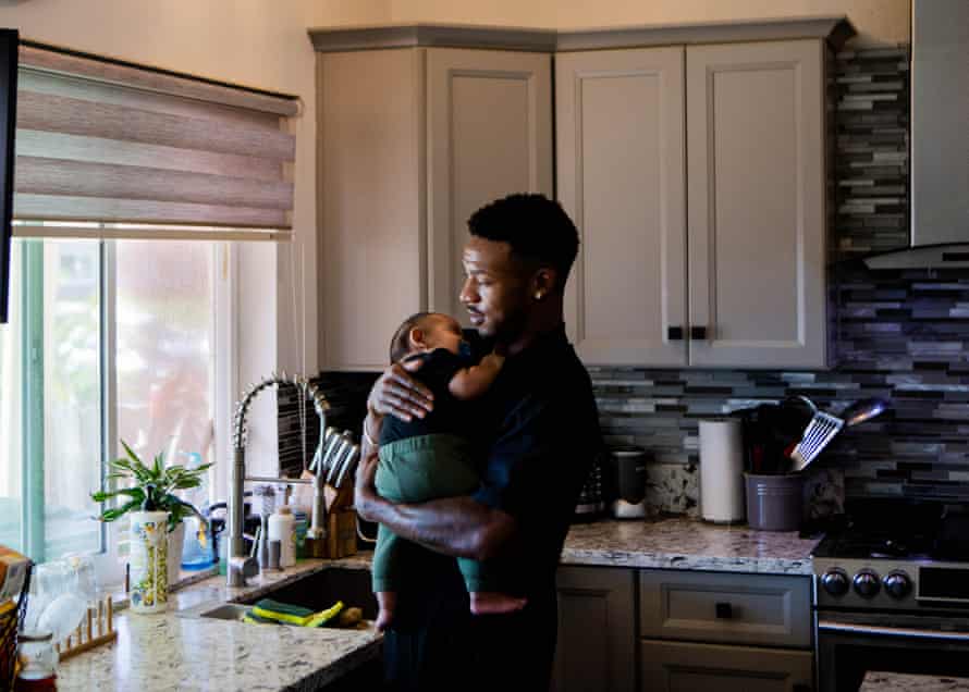 A black man holds his baby in a kitchen with gray cabinents