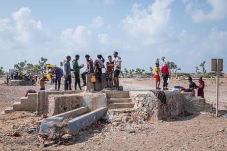 People stand on a concrete plinth reached by steps in the middle of an expanse of dirt and sand. A second water point with even more people can be seen a few metres away.
