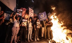 Israelis demonstrate in Tel Aviv demanding a prisoner swap deal and Netanyahu's resignation: they are holding placards including one saying Stop the War and standing behind a large fire in the street. It is dark and they are illuminated in the firelight.