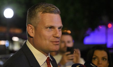 Close-up head and shoulders photo of Filip Turek: he has short, brushed-back blond hair and a square jaw, and wears a dark jacket, white shirt and red tie; it is dark and people are seen in the background taking photos or filming him on their phones
