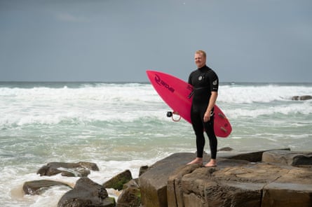 Jackson Baker stands on the rocks at the water’s edge on a beach, wearing a black wetsuit and holding his pink board