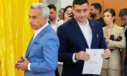 George Simion holds his ballot paper alongside Călin Georgescu, who is turned to face away from him. Both men wear white shirts with open necks; Georgescu is in a pale blue suit and Simion in a dark blue one.