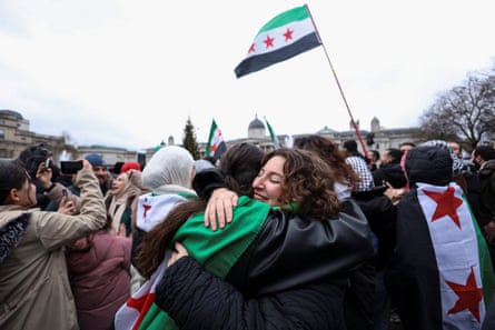Two young women embrace each other in a happy-looking crowd. People are waving Syrian opposition flags with one person wearing one across their shoulders.
