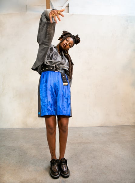 Little Simz standing with one arm up  and fingers down rapper-style, wearing big shiny blue shorts, silver grey top and chunky shoes