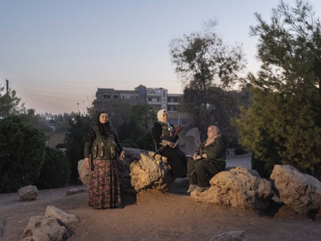 Two women sit on rocks outside in low light, a third stands beside them
