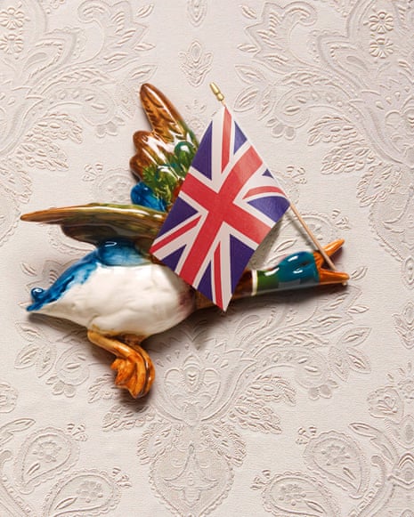 Three flying duck ornaments against a wallpapered wall, the front one carrying a union jack flag in its beak