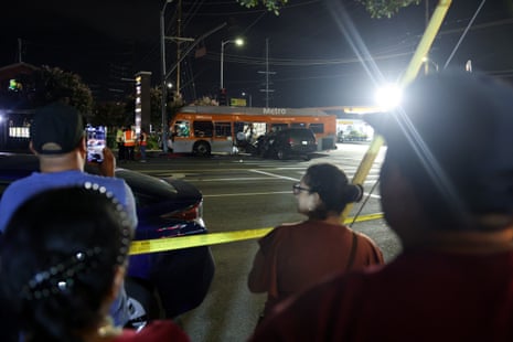 Bystanders watch a crashed LA Metro bus and mangled car behind police tape at night
