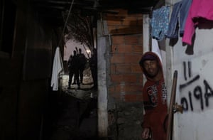 A night shot of a man in a hooded top looking out of doorway while behind him three heavily armed officers walk up an alleyway