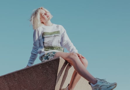 A woman with short bleached blond hair wearing a T-shirt and skirt and white trainers, with the blue sky behind her
