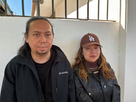 A man with long hair and a beard, wearing a dark anorak, and a woman with long, auburn hair wearing a brown LA baseball cap and a black jacket stand against a white wall.