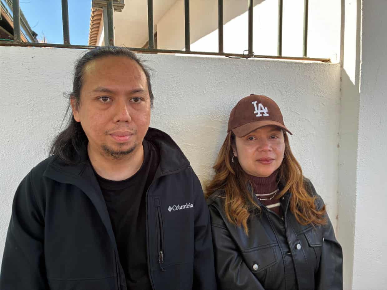 A man with long hair and a beard, wearing a dark anorak, and a woman with long, auburn hair wearing a brown LA baseball cap and a black jacket stand against a white wall.
