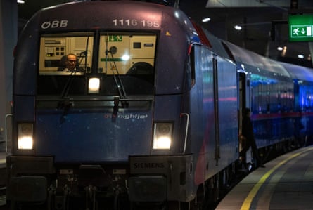 Front view of a dark blue Nightjet train with illuminated headlights as it waits to depart from Vienna; the driver's cabin is illuminated and a man sits at the controls.