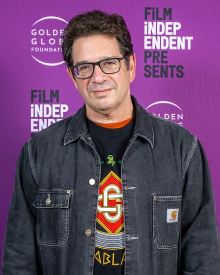 Peter Ettedgui attends a screening. He is casually dressed in a dark grey denim workwear jacket over a sweatshirt, and wears black-framed, square spectacles.