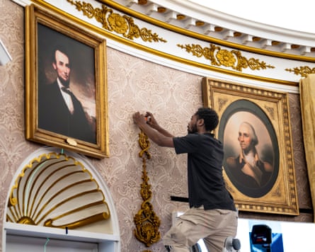 A man on a ladder glues ornate flourishes to a curved, wallpapered wall.