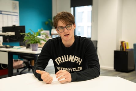 Charlie Pryce, 19, sits at a white table in a classroom-like space. He has brown hair with a long fringe and dark-rimmed glasses. He wears a black sweatshirt with the logo of Triumph Motorcycles.
