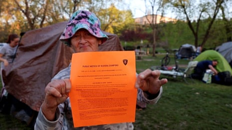 Weathered-looking white woman wearing pink and blue sunhat holds up bright orange piece of paper, with tents on green grass behind her.